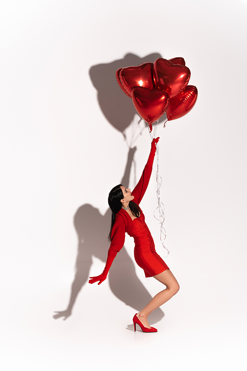 Side view of stylish woman in dress and heels pulling heart shaped balloons on white background with shadow