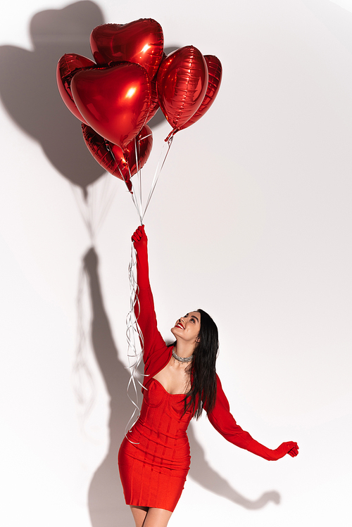 Cheerful woman in dress and gloves looking at red balloons in heart shape on white background with shadow