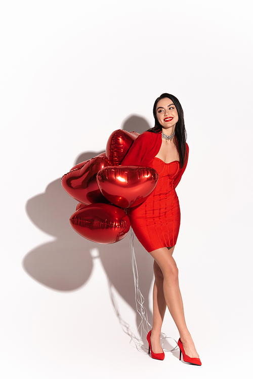 Stylish brunette woman in heels looking away near red heart shaped balloons on white background with shadow