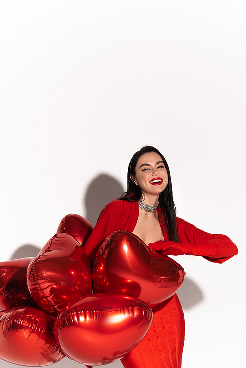 Stylish woman with red lips looking at camera near heart shaped balloons on white background with shadow