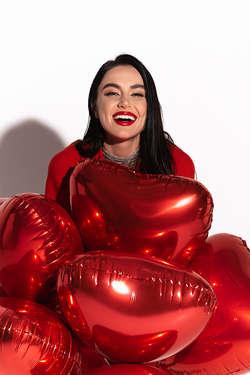 Positive brunette model with red lips looking at camera near heart shaped balloons on white background with shadow