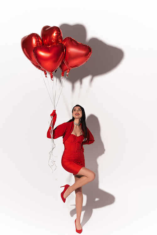 Stylish woman in red dress and heels looking at balloons in heart shape on white background with shadow