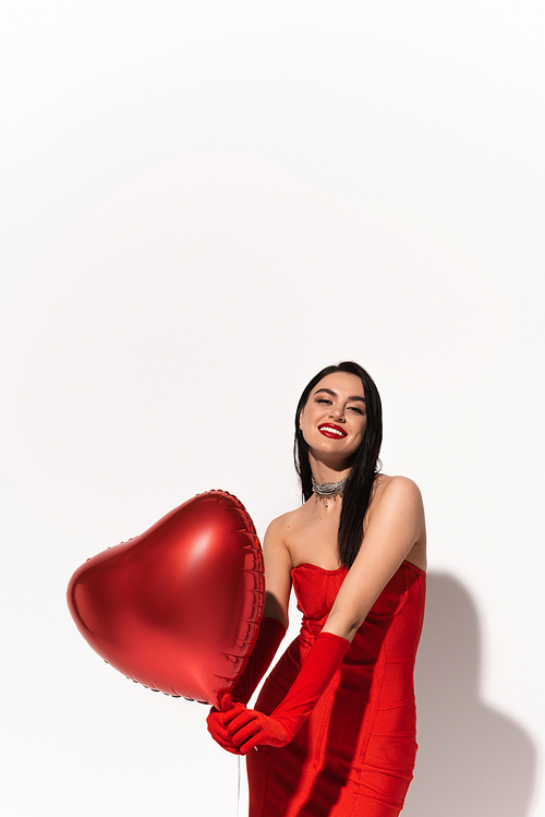 Smiling woman in red dress holding heart shaped balloons and looking at camera on white background with shadow
