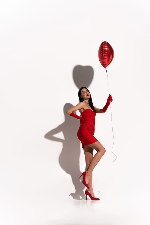Full length of sexy woman in red dress and gloves holding heart shaped balloon on white background with shadow