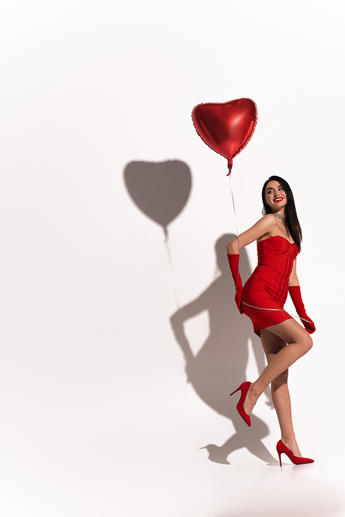 Sexy brunette woman in heels and red gloves holding heart shaped balloon on white background with shadow
