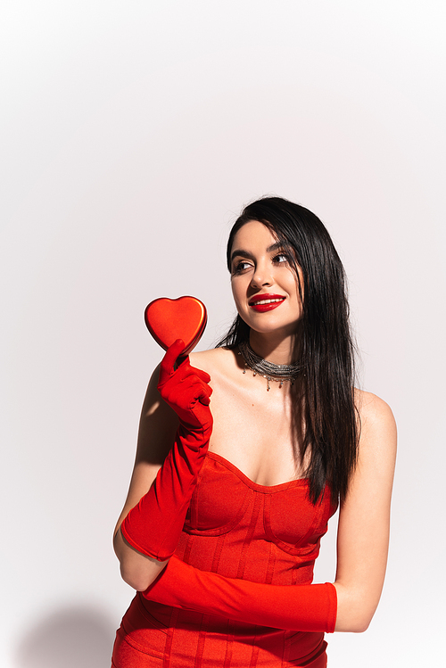 Stylish woman in red dress and gloves holding gift box in heart shape on grey background