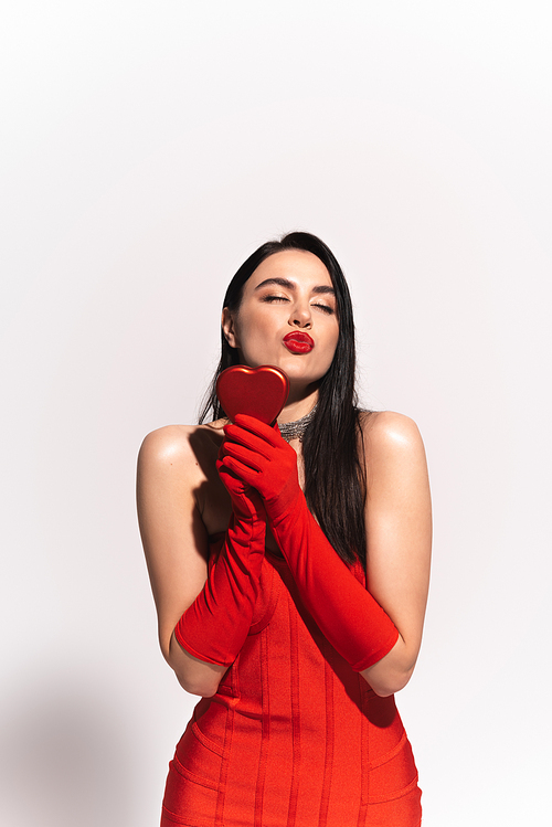Stylish model in red dress and gloves holding heart shaped gift box and blowing air kiss on grey background