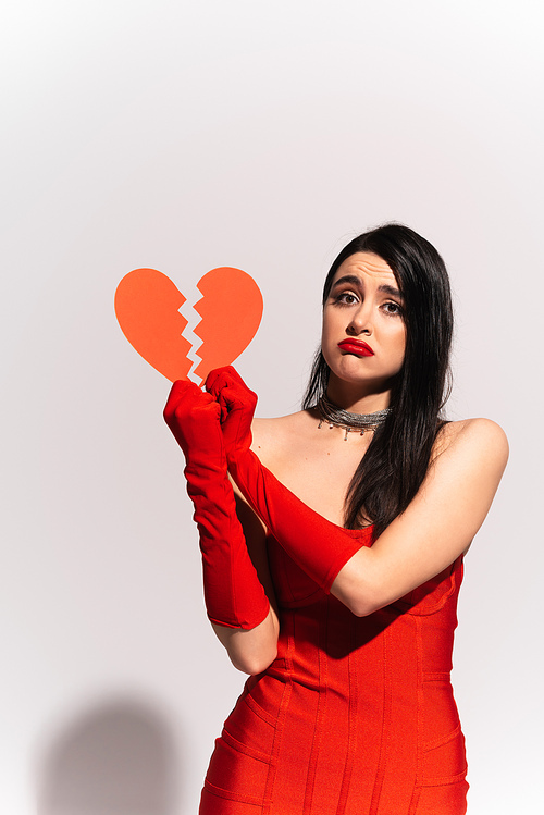 Displeased woman in red gloves and dress holding broken paper heart on grey background
