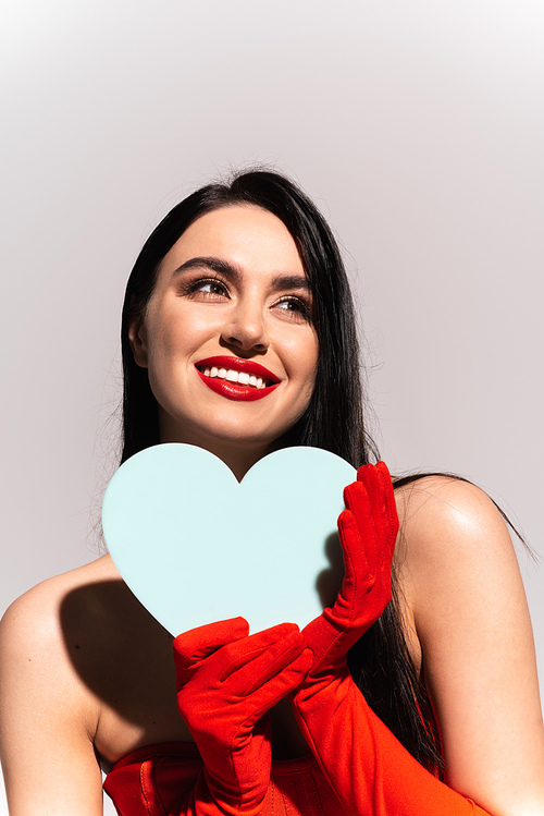 Smiling brunette woman in red gloves holding paper heart isolated on grey