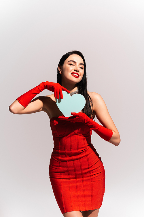 Pleased woman in red dress holding paper heart isolated on grey