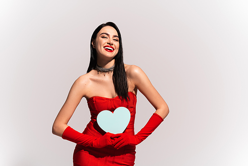 Positive model in red dress and gloves holding paper heart isolated on grey