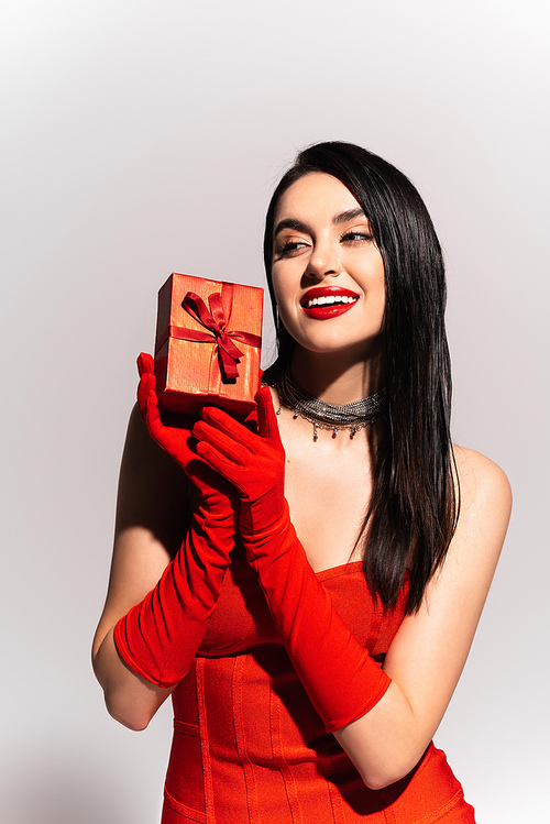 Stylish brunette woman in red gloves holding gift box on grey background