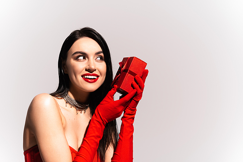Cheerful brunette woman in red gloves holding present isolated on grey