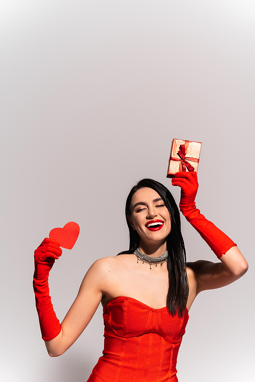 Cheerful brunette woman in red gloves holding gift and paper heart isolated on grey