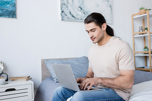 freelancer with long hair sitting on bed and using laptop