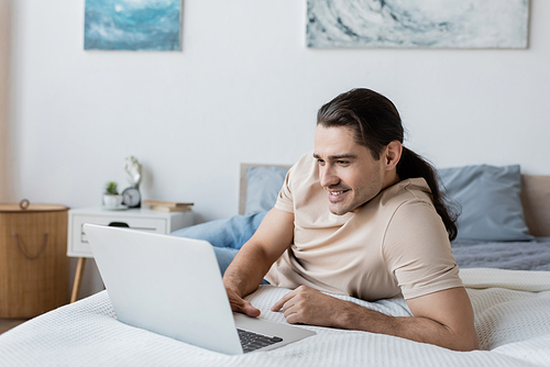 cheerful freelancer with long hair using laptop in bedroom