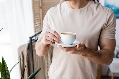 cropped view of man in t-shirt holding saucer with cup of coffee