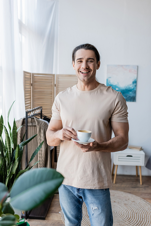 cheerful man holding cup of coffee and saucer near green plants