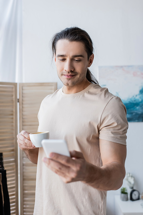 man in t-shirt holding cup of coffee and using smartphone