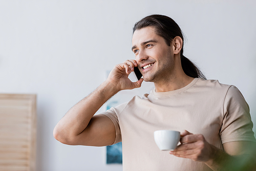 happy man with long hair holding cup of coffee and talking on smartphone