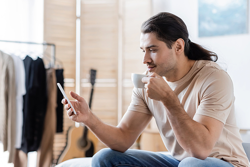 man with long hair holding cup of coffee and using mobile phone