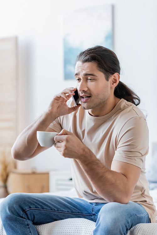 man with long hair holding cup of coffee and talking on mobile phone