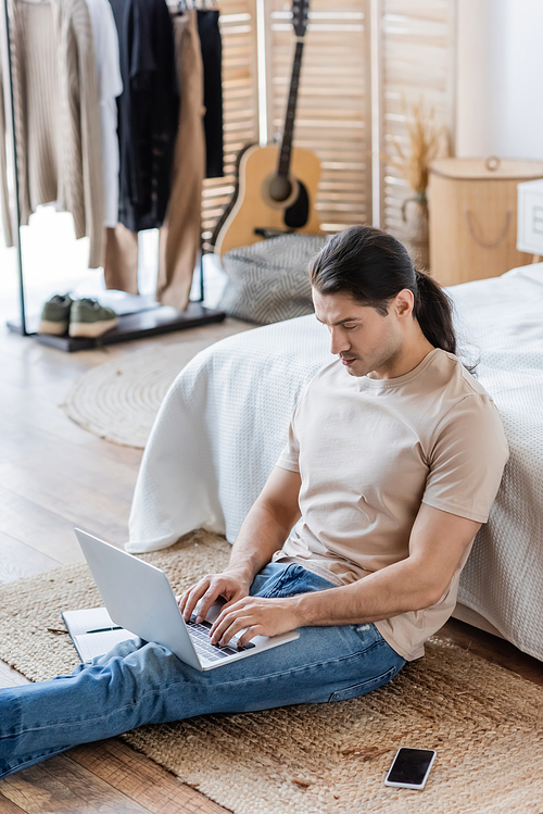 man with long hair using laptop while sitting on floor in bedroom