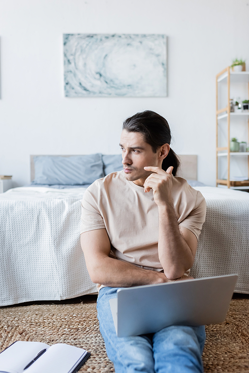 pensive freelancer looking away near laptop and notebook in bedroom