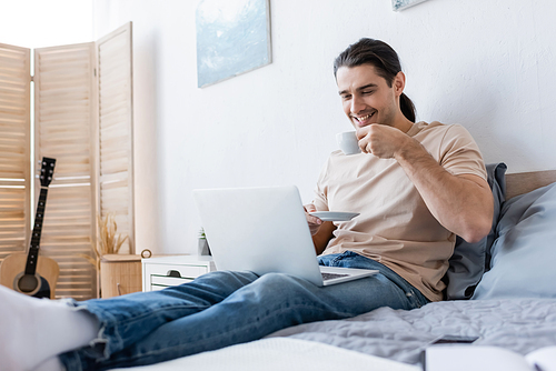 cheerful man with long hair holding cup of coffee while using laptop in bedroom