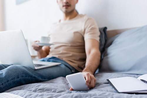 cropped view of blurred man holding cup of coffee while reaching smartphone in bedroom