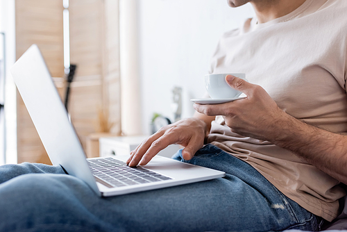 cropped view of man holding cup of coffee while using laptop in bedroom