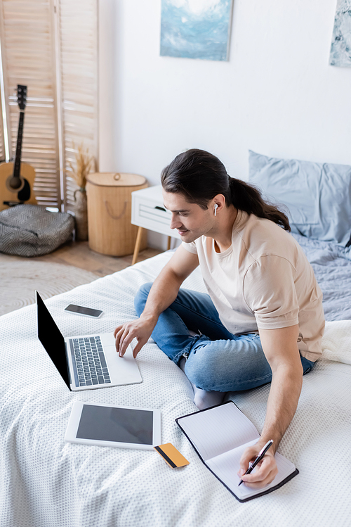 smiling man with long hair in earphone writing in notebook and using laptop near gadgets on bed