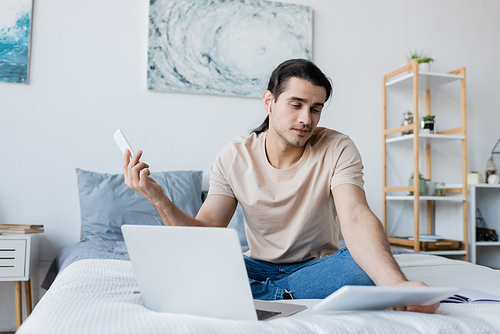 man in earphone holding smartphone while looking at digital tablet near laptop on bed