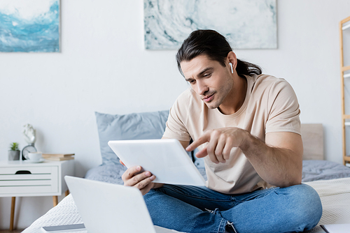 man in earphone pointing at digital tablet near laptop and smartphone on bed