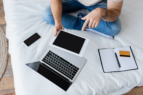 cropped view of man pointing at digital tablet with blank screen near laptop and smartphone on bed
