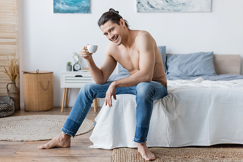 cheerful man with long hair holding cup of coffee while sitting on bed