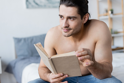 dreamy and shirtless man with long hair holding book in bedroom