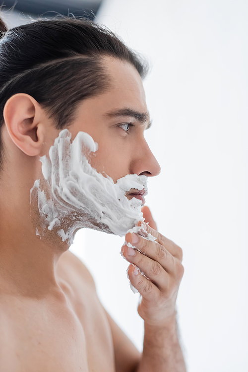 side view of shirtless man applying shaving foam on face in bathroom