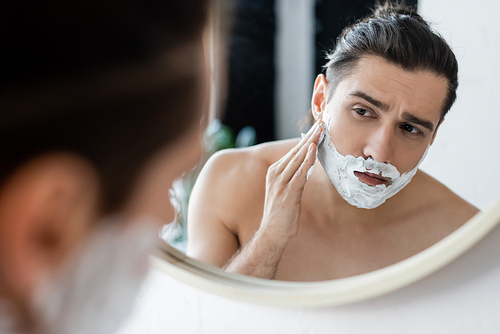 shirtless man applying shaving foam on face and looking at mirror