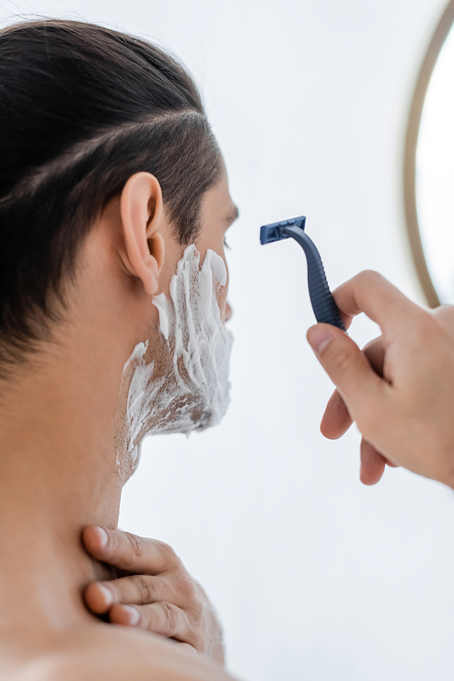 shirtless man with shaving foam on face holding safety razor in bathroom