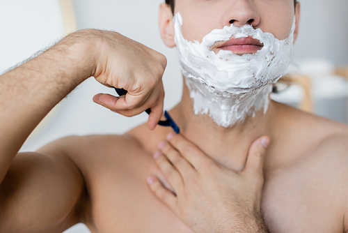 partial view of shirtless man with foam on face shaving in bathroom