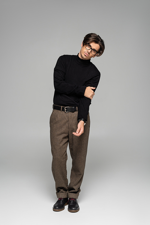 full length of brunette man in black turtleneck and pants posing in eyeglasses on grey