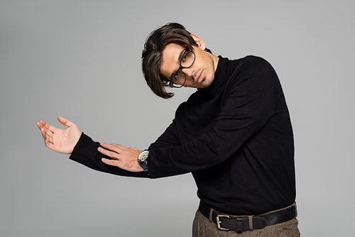 young man in black turtleneck and eyeglasses looking at camera while posing isolated on grey