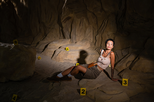 young brunette archaeologist sitting near numbered cards in dark cave and looking away