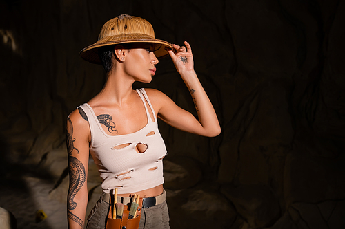 tattooed archaeologist with tools in waist bag adjusting safari hat while looking away in dark desert