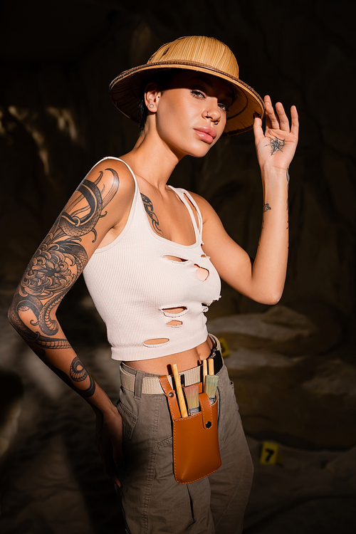sexy tattooed archaeologist in tank top touching safari hat and looking at camera in dark cave