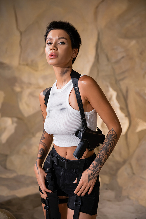 tattooed young archaeologist with dirty crop top standing with gun in holster