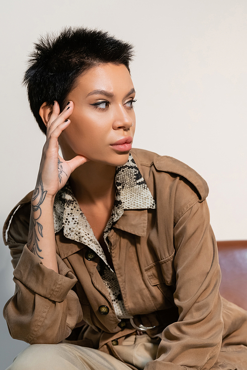 brunette archaeologist in beige jacket with tattooed hand near face looking away on grey