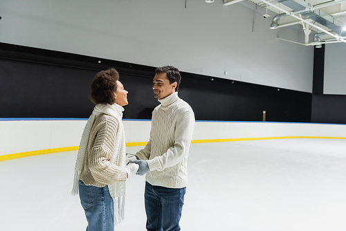 Multiethnic couple in warm knitted sweaters holding hands on ice rink