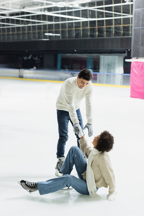 Young man giving hand to african american girlfriend falling on ice rink